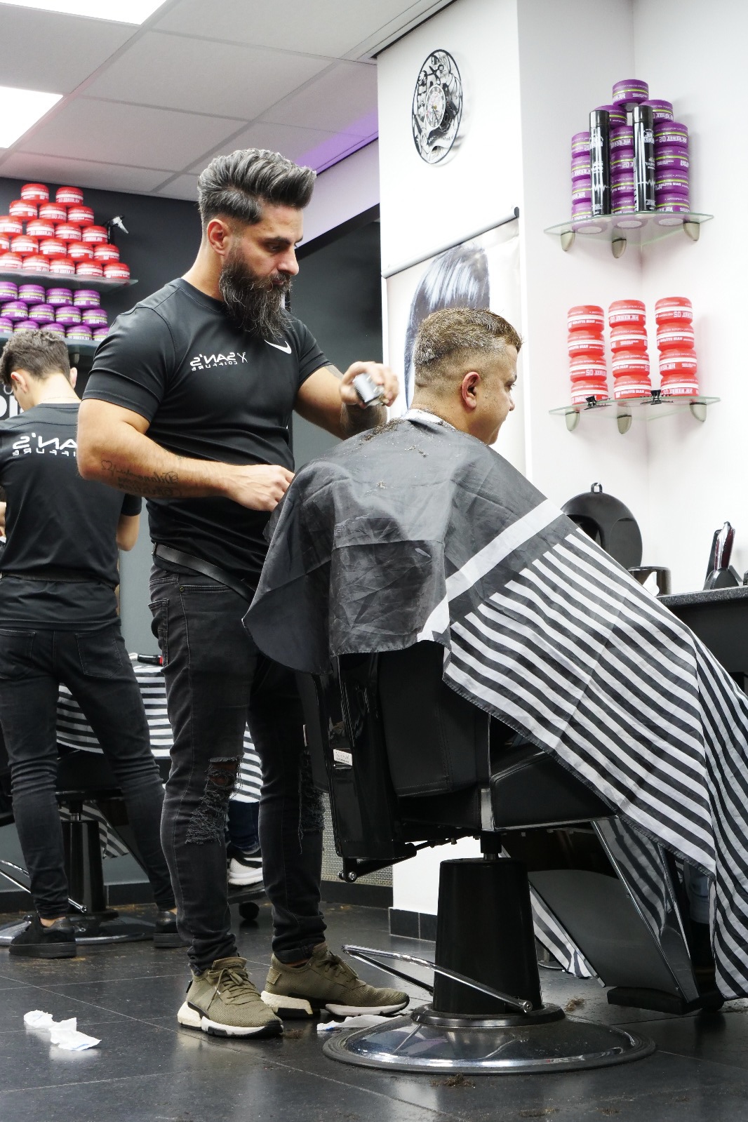 Een barber van San’s Coiffure knipt een klant in een gestreepte kapmantel, met de wax-wand op de achtergrond
