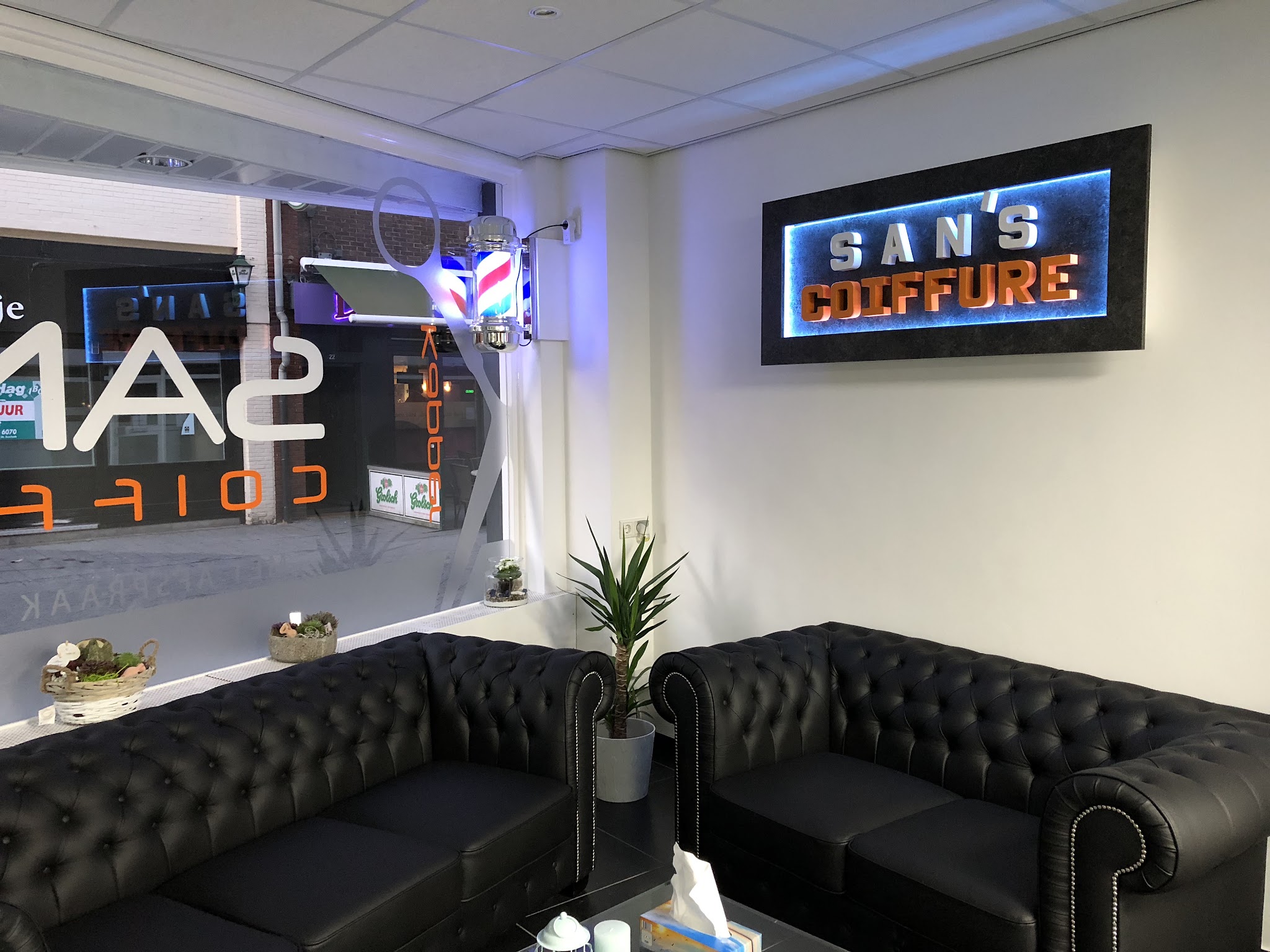 De wachtruimte met zwarte chesterfield-banken en het verlichte San’s Coiffure bord