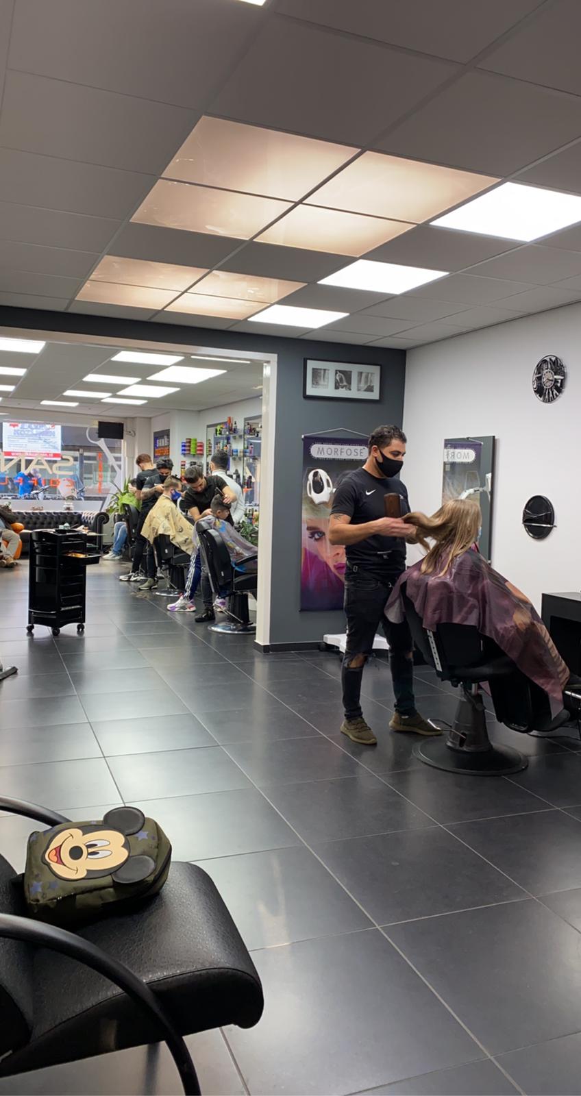 Een lange rij stoelen met barbers aan het werk en klanten in kapmantels